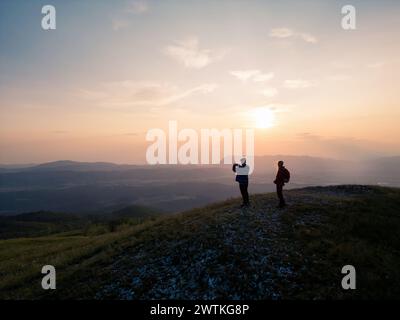 Ragazzo adolescente che scatta foto del fantastico paesaggio montano al tramonto con lo smartphone mentre si trova in un punto panoramico in compagnia di una ragazza con lo zaino. Foto Stock