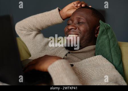 Ritratto di un uomo afroamericano sorridente sdraiato sul divano e che guarda lo schermo del laptop mentre si rilassa al lavoro Foto Stock