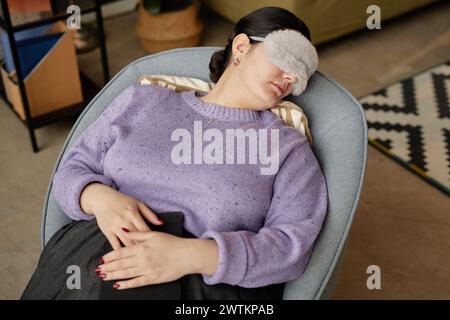 Vista ad alto angolo su una giovane donna che dorme in poltrona al lavoro con la maschera di sonno attivata Foto Stock