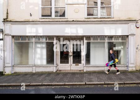 Una giovane ragazza che porta con sé uno skateboard colorato e passa davanti a un negozio chiuso vuoto in Fore Street nel centro di Bodmin in Cornovaglia Foto Stock