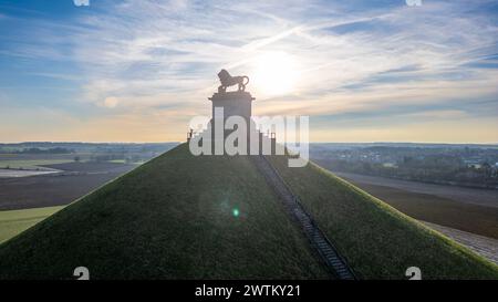 Waterloo, Bruxelles, Belgio, 25 febbraio 2024, si tratta di una tranquilla fotografia all'alba del tumulo dei leoni presso il memoriale del campo di battaglia di Waterloo. Il sole nascente crea un alone luminoso intorno alla statua del leone, che si erge in cima al piedistallo in pietra, creando una silhouette impressionante. I raggi del sole si riversano sul tumulo e sulla campagna circostante, illuminando il sentiero che serpeggia sulla collina. Gli ampi campi sullo sfondo sono immersi nella luce del mattino presto, dando al paesaggio una qualità tranquilla ed eterea. Lion's Mound Memorial silhouette al Sunris. Foto di alta qualità Foto Stock