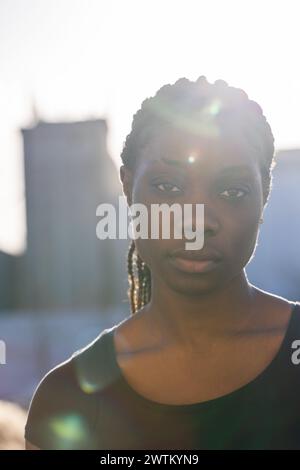 Una donna africana è retroilluminata dal sole che tramonta, la sua silhouette è parzialmente delineata contro il soffice bagliore del cielo serale. I raggi del sole filtrano i suoi capelli, creando un effetto alone naturale intorno alla sua testa. La sua espressione è solenne e introspettiva, con lo skyline della città che forma uno sfondo silenzioso. La calda retroilluminazione e il leggero bagliore dell'obiettivo aggiungono una qualità onirica e riflettente all'immagine. Donna africana retroilluminata con silhouette urbana. Foto di alta qualità Foto Stock