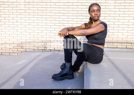 Una donna afroamericana siede con fiducia a terra, la schiena contro un muro di mattoni bianchi. Sfoggia un outfit casual nero abbinato a stivali da combattimento che incarnano uno stile urbano. Le sue trecce e l'espressione contemplativa aggiungono profondità al suo comportamento casuale ma determinato. L'illuminazione è luminosa e proietta la sua ombra sul terreno, il che aggiunge all'estetica urbana dell'immagine. Donna elegante seduta contro Brick Wall. Foto di alta qualità Foto Stock