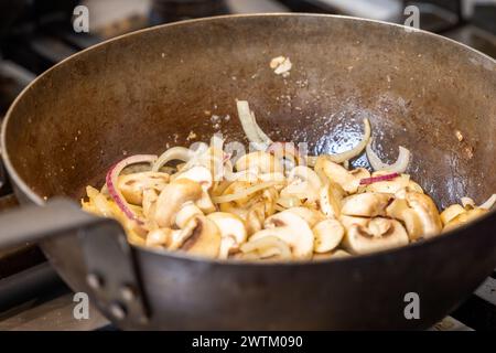 L'immagine cattura una vista ravvicinata di funghi e cipolle appena affettati mentre sorseggiano in una padella ben condita, che suggerisce molti usi precedenti e pasti saporiti. Il vapore aumenta leggermente, indicando il calore del processo di saltatura. Le cipolle iniziano a diventare traslucide e i funghi leggermente marroni, a indicare il perfetto stadio di cottura. Questo momento culinario è un passo comune ma essenziale in molte ricette, promettendo un delizioso contributo al piatto finale. Affettare funghi e cipolle in una padella calda. Foto di alta qualità Foto Stock