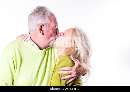 Questa immagine avvolgente cattura un momento tenero tra una coppia senior mentre condividono un bacio amorevole. Entrambi sono vestiti con maglioni vivaci, con uno sfondo bianco che accentua l'intimità e il calore dell'abbraccio. Affettuosa coppia senior che condivide un bacio. Foto di alta qualità Foto Stock