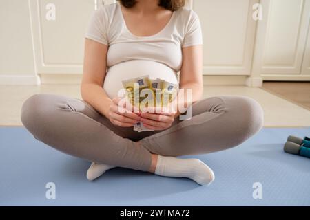 Corsi di fitness durante la gravidanza e spese finanziarie. Donna incinta ad una sessione di allenamento sportivo in cucina con soldi in euro in mano, prepa Foto Stock