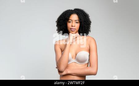 Una donna afro-americana con un sorriso radioso e ricci naturali galleggianti si mantiene sicura. Le mani sui fianchi e la posa spensierata su uno sfondo grigio creano un'immagine di gioiosa fiducia. Foto Stock