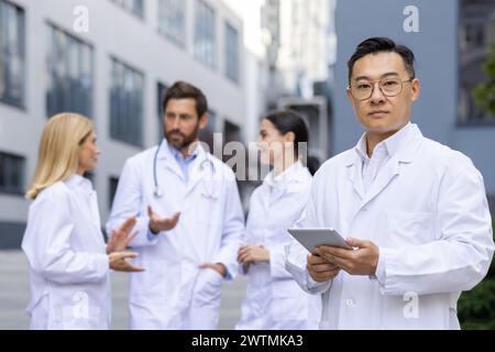 Medico cinese serio che guarda la fotocamera e tiene in mano un tablet digitale mentre si sta in piedi su uno sfondo sfocato con i colleghi. Gruppo di operatori medici in mentre camici di laboratorio discutere nuova strategia di trattamento. Foto Stock