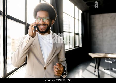 Un vivace uomo d'affari brasiliano è impegnato in una conversazione telefonica in un moderno loft Office, con luce del sole che scorre attraverso grandi finestre, catturando un momento di vita aziendale dinamica Foto Stock