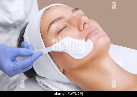 Il cosmetologo per la procedura di pulizia e idratazione della pelle, applicando una maschera alginica al viso di una giovane donna nel salone di bellezza.Cosmetolo Foto Stock