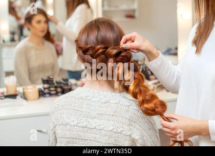 Bella ragazza, con lunghi capelli rossi. Il parrucchiere intreccia una treccia francese, primo piano in un salone di bellezza. Cura professionale dei capelli e creazione dei capelli Foto Stock