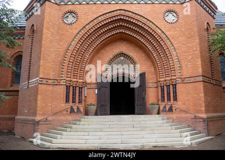 Ingresso principale all'edificio di una chiesa neogotica di Visitazione della Vergine Maria, fatta di mattoni arancioni. Cancello di legno marrone scuro aperto di grandi dimensioni. Postorna Foto Stock