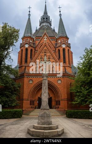 Costruzione di una chiesa neogotica di Visitazione della Vergine Maria, fatta di mattoni arancioni. Statua della Crocifissione nel parco. Postorna, Breclav, Moravia, CZ Foto Stock