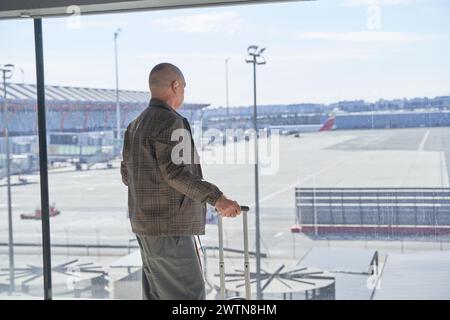 Un tranquillo turista di sesso maschile è in piedi in aeroporto e guarda il volo degli aerei attraverso la finestra. Foto Stock