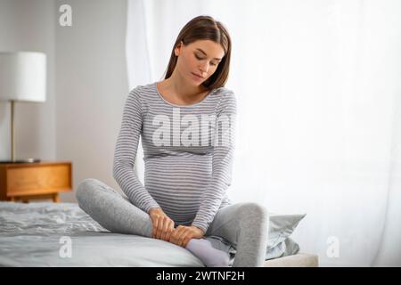 Giovane donna incinta in abbigliamento casual che massaggia la caviglia gonfia a casa Foto Stock