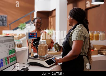 Una donna nera in grembiule assiste un cliente al banco del negozio ecologico con pesatura di prodotti biologici locali. Un giovane cliente che tiene in mano una borsa della spesa che si avvicina alla cassa mentre il venditore utilizza una bilancia digitale. Foto Stock