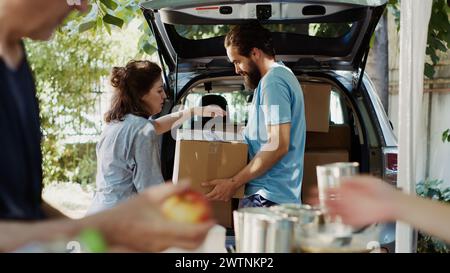 Durante la guida alimentare, il volontario maschile dà scatole di donazione dall'auto agli individui bisognosi e meno privilegiati. Gruppo di volontari che distribuiscono le necessità ai poveri e ai senzatetto. Scatto con cavalletto. Foto Stock