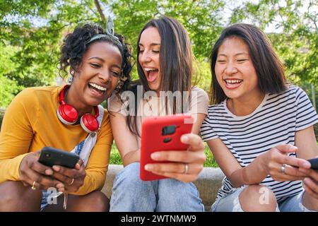 Tre gioiose donne multirazziali che usano il telefono seduto all'aperto in estate. Giovane donna sorprende ridendo Foto Stock