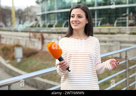 Giovane giornalista femminile con microfono che lavora per la strada cittadina Foto Stock