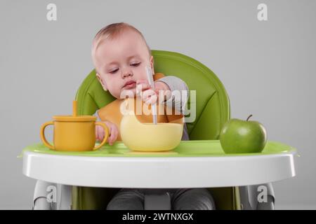 Piccolo bambino carino che mangia cibo sano in seggiolone su sfondo grigio Foto Stock