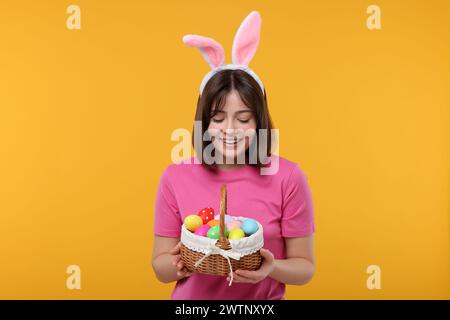 Festa di Pasqua. Donna felice con orecchie conigliette e cesto di vimini pieno di uova dipinte su sfondo arancione Foto Stock