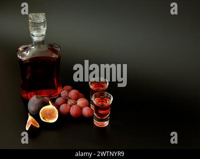 Due bicchieri e un decanter in vetro con liquore fatto in casa su sfondo nero, accanto a pezzi di fichi maturi e un mazzo di uva rossa. Primo piano. Foto Stock