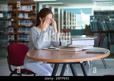 Giovane studentessa asiatica che sfrega gli occhi, si sente stanca dopo aver letto un libro in biblioteca. Foto Stock