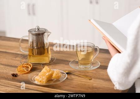 Composizione con teiera, tazza di tè e una giovane donna che legge un libro in cucina Foto Stock