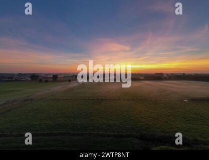 Questa fotografia grandangolare cattura la bellezza tranquilla di un'alba che diffonde il suo caldo bagliore su un pascolo lussureggiante. L'orizzonte è adornato da una serie di colori, che vanno dai viola profondi alle arance ardenti e ai gialli morbidi, mentre il sole sbircia sopra di esso, suggerendo la rottura dell'alba. La luce precoce proietta ombre delicate ed evidenzia le texture del campo e la linea della recinzione. In lontananza, sono visibili sottili accenni di civiltà, possibilmente una piccola comunità o case rurali, abbracciate dal cielo che si risveglia. L'immagine evoca un senso di pace e la semplice grandezza dello spettacolo quotidiano della natura. S Foto Stock