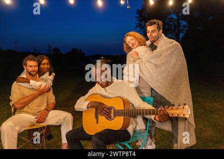 Un gruppo di amici gode di un momento sereno sotto il cielo notturno, avvolto da coperte, mentre si suona la chitarra, creando un'esperienza all'aperto accogliente e musicale. Accogliente serenata notturna presso un raduno all'aperto. Foto di alta qualità Foto Stock