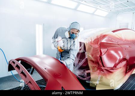 Pittore di auto in indumenti protettivi e maschere di verniciatura paraurti auto con vernice rossa e vernice in officina camera. Foto Stock