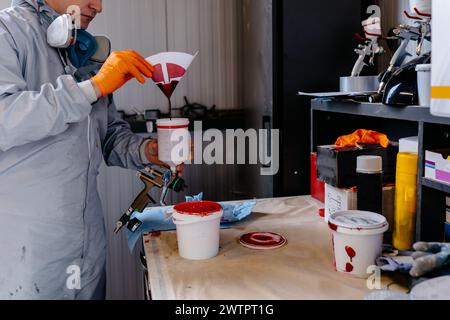 Verniciatore di auto con indumenti protettivi che versano vernice mista da una lattina in un secchio di miscelazione in plastica presso la stazione di servizio dell'auto. Foto Stock