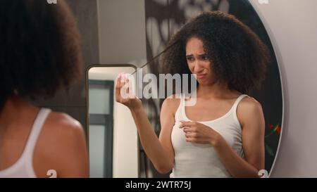 Frustrata esausta pazza arrabbiata triste donna afroamericana che cerca di pettinare i ricci soffre con problemi di cura dei capelli guardando specchio riflesso ragazza femmina Foto Stock