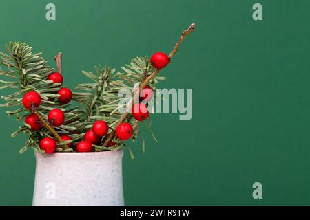 Bouquet di nobilis abete e rami di winterberry in un vaso di fronte allo sfondo verde. Concetto con spazio di copia. Foto Stock