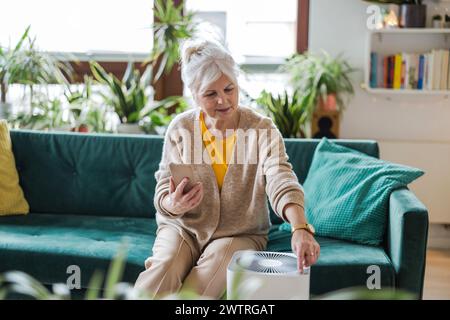 Donna anziana che utilizza lo smartphone per configurare un purificatore d'aria domestico Foto Stock