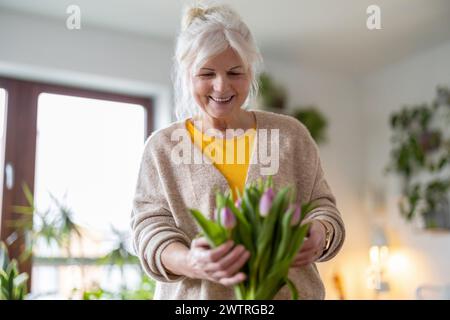 Donna anziana felice con bouquet di tulipani in vaso a casa Foto Stock