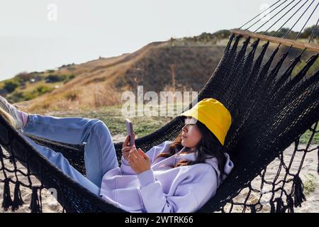 Una giovane e attraente donna asiatica tiene un telefono cellulare e giace in un'amaca. Un'affascinante donna coreana in abiti casual usa uno smartphone e si rilassa Foto Stock