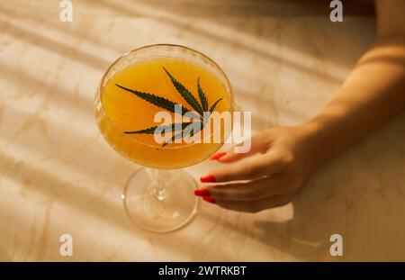 Una mano femminile con una bella manicure rossa raggiunge un elegante bicchiere a gambo lungo con un cocktail con foglie di cannabis. Una deliziosa bevanda gialla con h Foto Stock