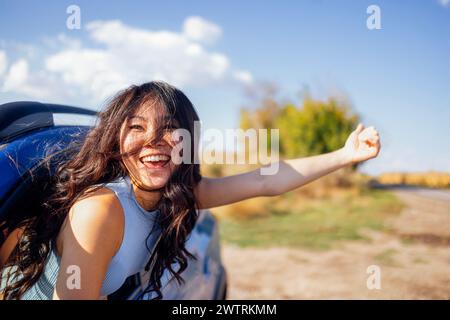 Una giovane asiatica si sporge dal finestrino dell'auto e ride. Una donna coreana sorridente viaggia e si diverte in viaggio. Una ragazza attraente si diverte a guidare e a Foto Stock