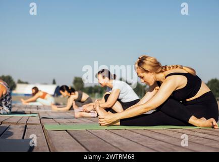 Lezione di yoga mattutina con istruttore vicino al fiume in città. Le giovani donne si piegano mentre si siedono sui tappetini in un molo di legno. Un gruppo di ragazze multietniche sono in agguato Foto Stock