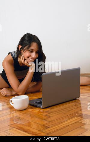 Immagine verticale di una giovane donna bruna ispanica latina con capelli neri e abbigliamento sportivo, seduta sul pavimento, felice, che tiene lezioni di yoga con il laptop a hom Foto Stock