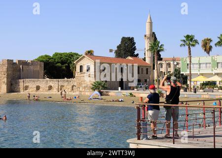 Coppia di turisti che scatta foto al forte di Larnaca e al minareto della Moschea. Bagnanti sulla spiaggia e nuotatori. Finikoudes, Larnaca, Cipro Foto Stock