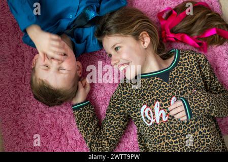 Due bambini che condividono un momento di gioia sdraiati su un tappeto rosa fuzzy Foto Stock