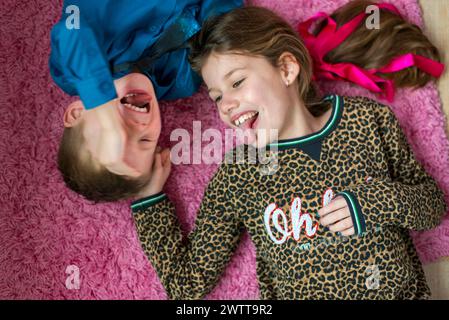 Due bambini che fanno facce divertenti mentre sdraiati su un tappeto rosa. Foto Stock