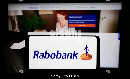 Persona che detiene un cellulare con il logo della società bancaria olandese Cooperatieve Rabobank U.A., di fronte alla pagina web aziendale. Mettere a fuoco il display del telefono. Foto Stock