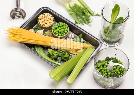 Pasta, verdure e anacardi in pallet. Insalata Romaine, piselli verdi e broccoli nel recipiente del frullatore. Vista dall'alto. Sfondo bianco. Foto Stock
