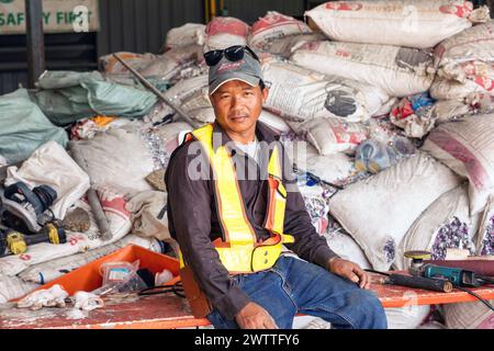 Giovane lavoratore in giubbotto riflettente seduto in mezzo a materiali industriali in una fabbrica di riciclaggio di plastica, in Thailandia Foto Stock