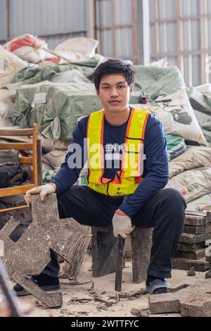 Giovane lavoratore in giubbotto riflettente seduto in mezzo a materiali industriali in una fabbrica di riciclaggio di plastica, in Thailandia Foto Stock