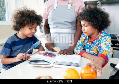 Bambini che leggono un libro in cucina con supervisione di un adulto Foto Stock