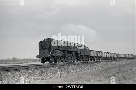 1960s, Historical, una locomotiva a vapore della British Railway 92216, su binari ferroviari che tirano carri merci, Inghilterra, Regno Unito. Con un design distintivo, le locomotive a vapore BR Classe 9F furono utilizzate per trainare carri merci pesanti, a velocità sostenuta, su lunghe distanze, ma furono utilizzate anche sui treni passeggeri. L'ultima locomotiva a vapore costruita dalla British Railways nel 1960 era una classe 9F, la 92220 Evening Star. Foto Stock